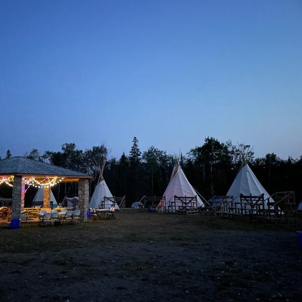 An outdoor scene at dusk with a stone pavilion and several tipis arranged in a circle with bleachers.